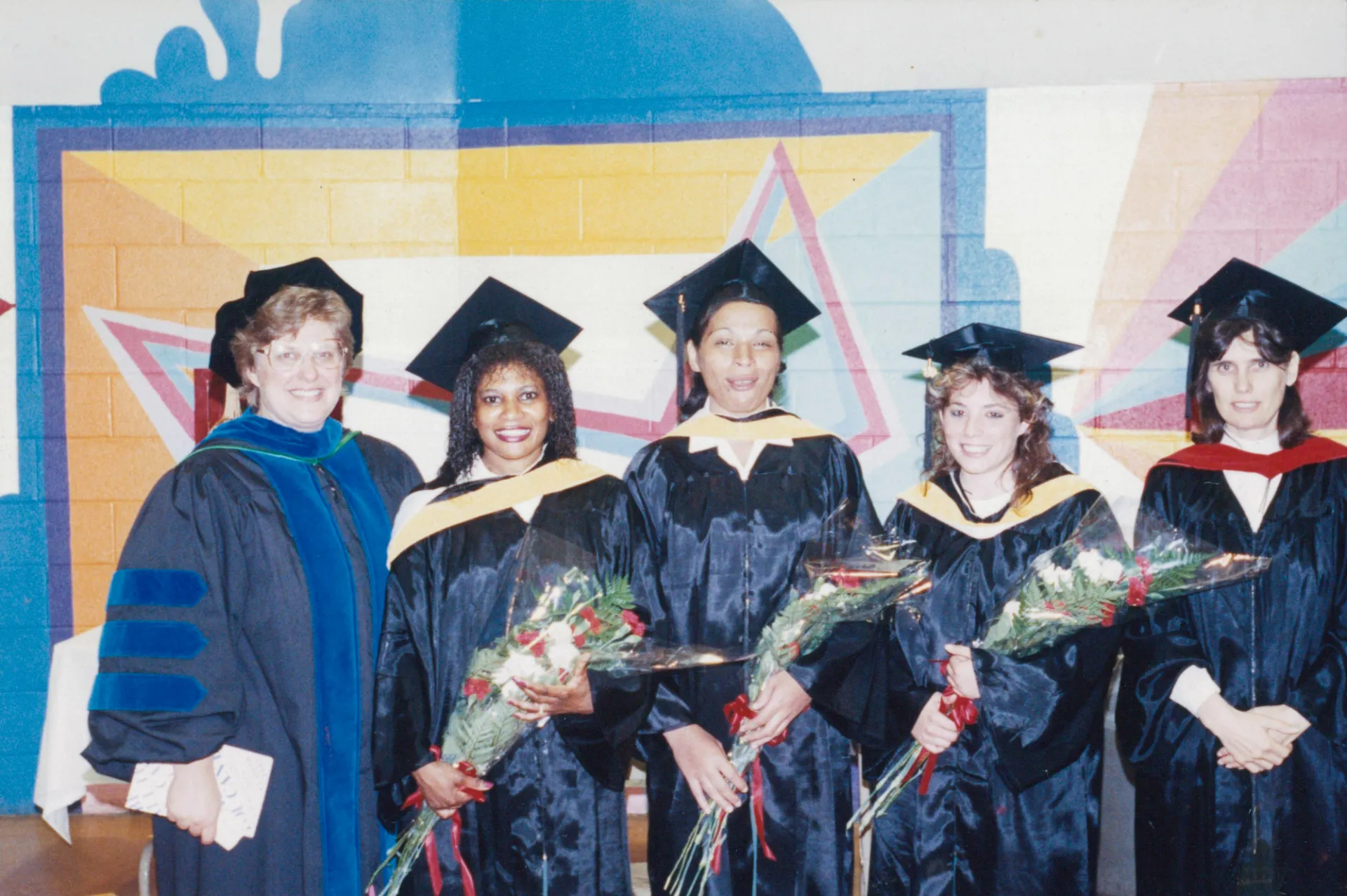 A group of women in caps and gowns holding flowers in front of a colorful background