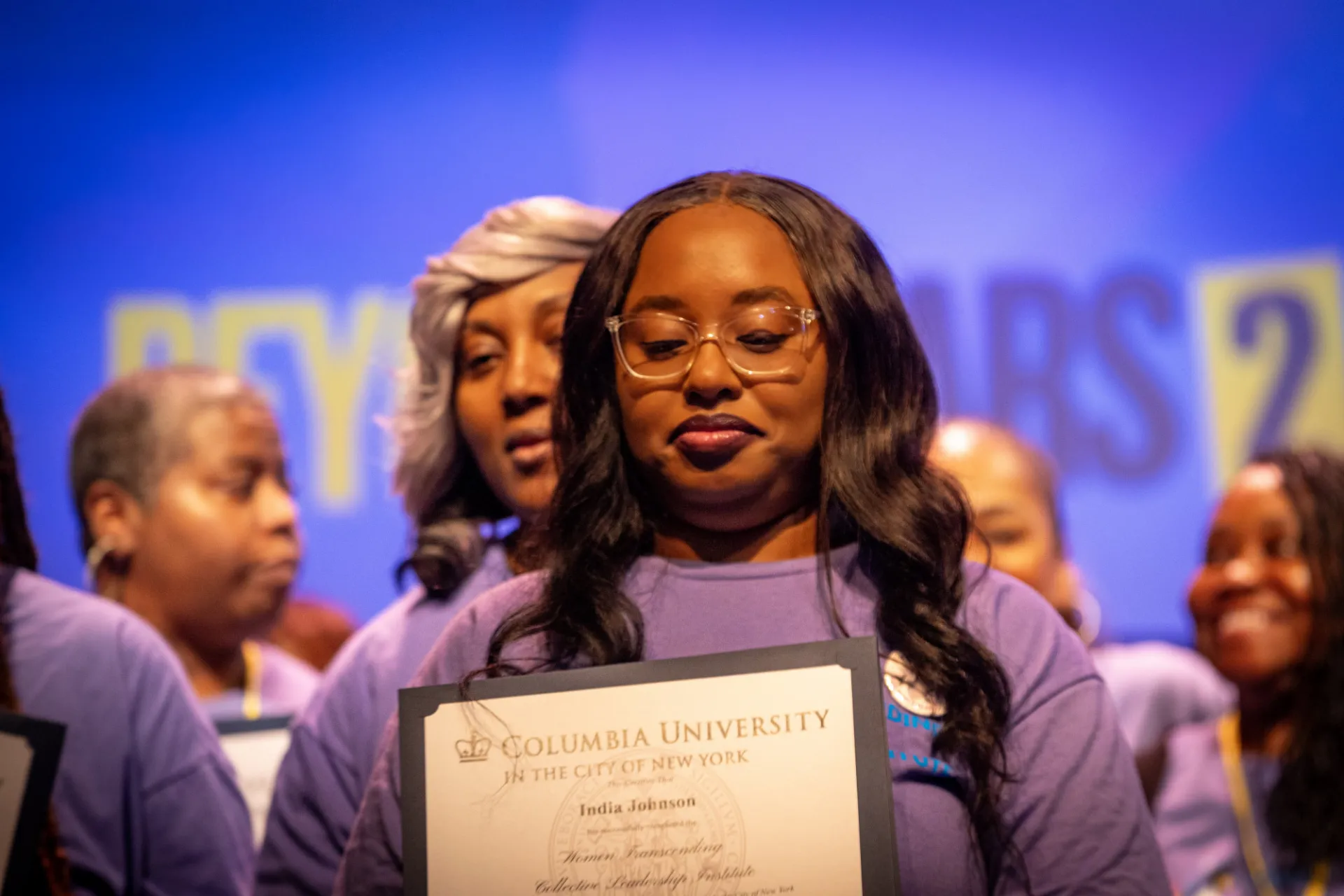 A woman in a purple shirt looking down and smiling and holding a certificate that says "Columbia University in the City of New York"