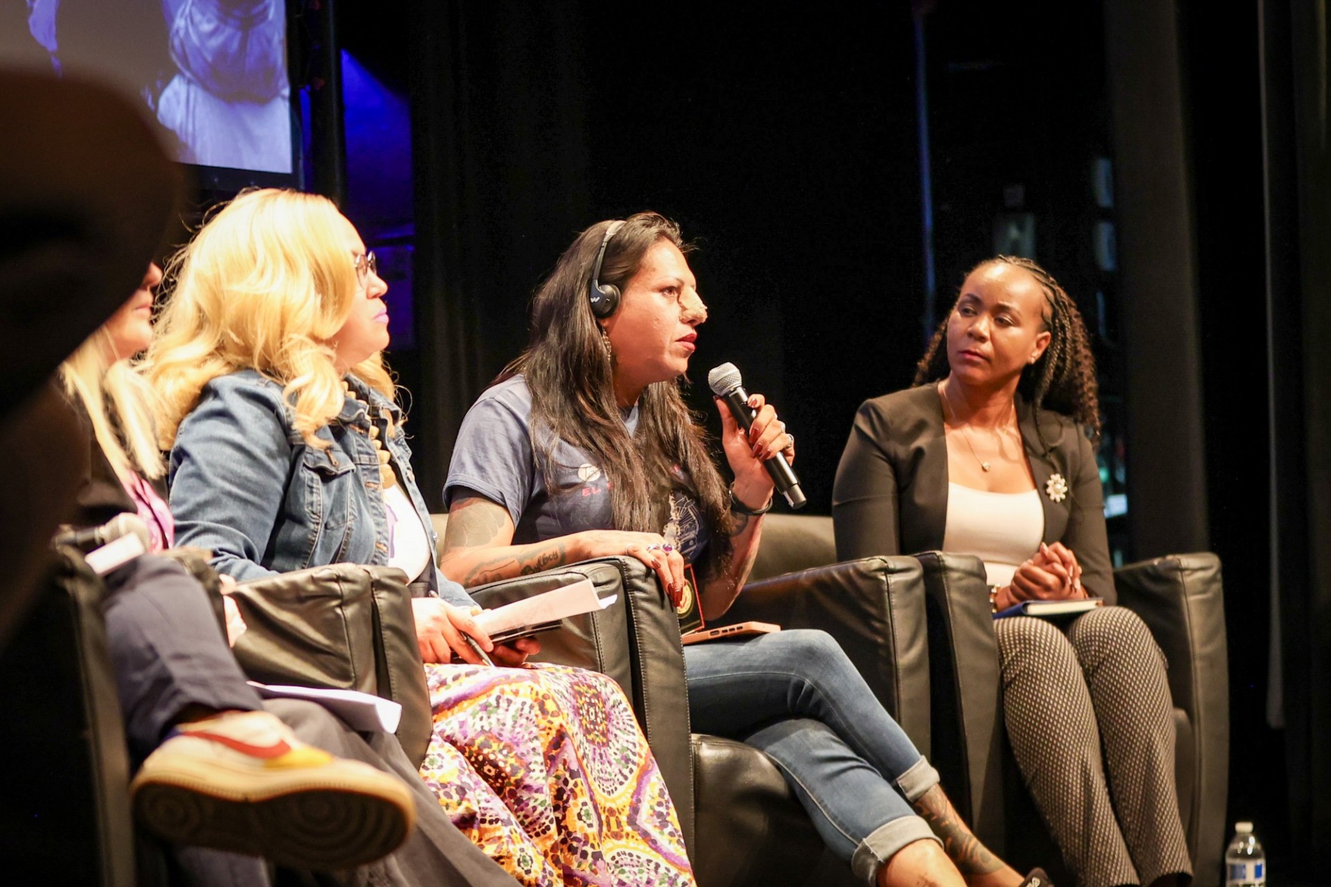 Panelists on stage in chairs with one holding a microphone