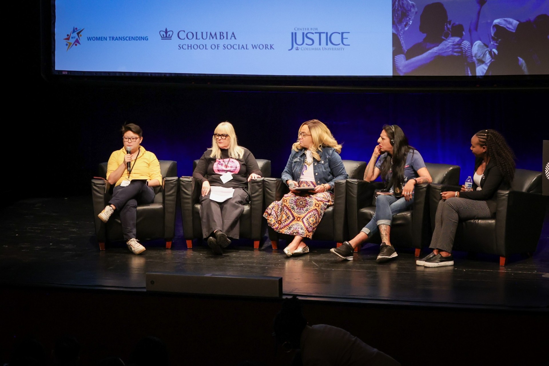 Five panelists sitting in chairs on a stage with the one on the far lieft holding a microphone