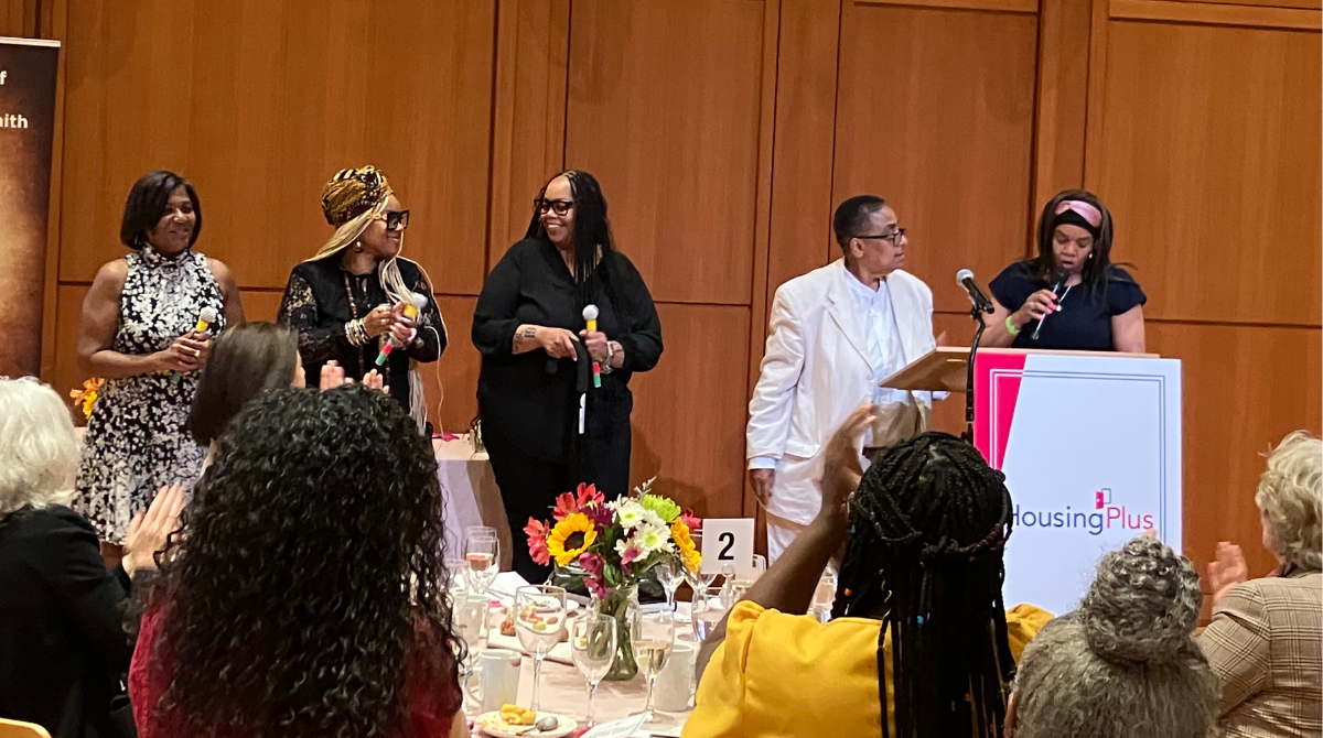 five Black women standing in front of an audience holding awards and microphones