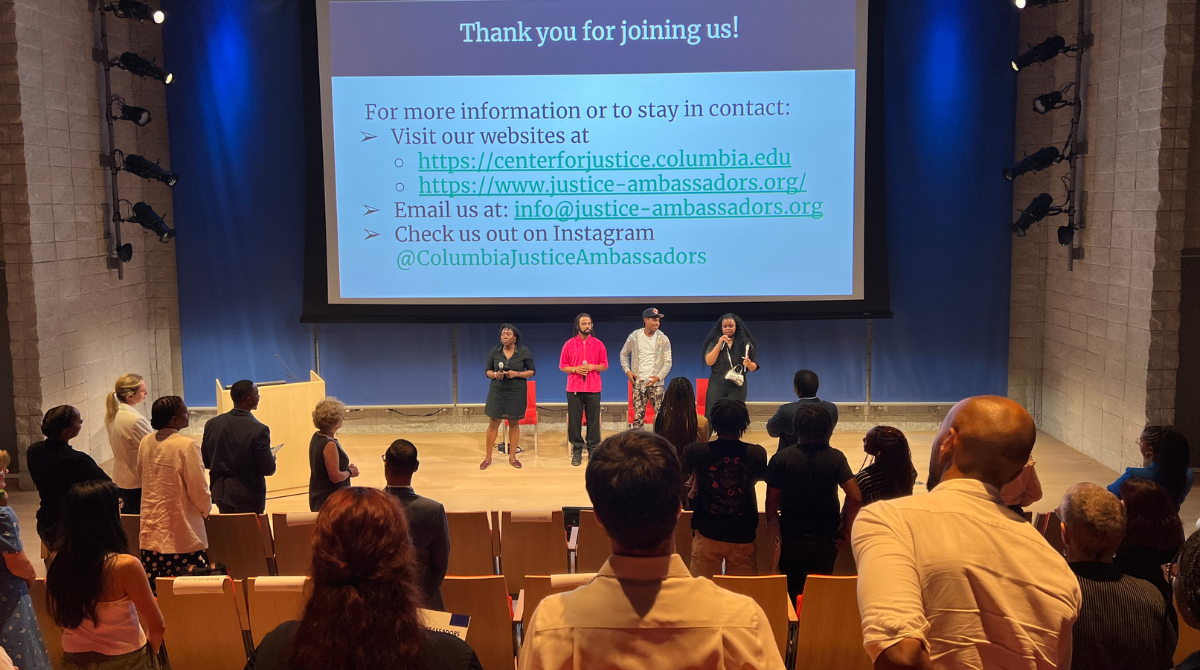 Photo of an auditorium with four people standing up front with mics and people in the audience standing 