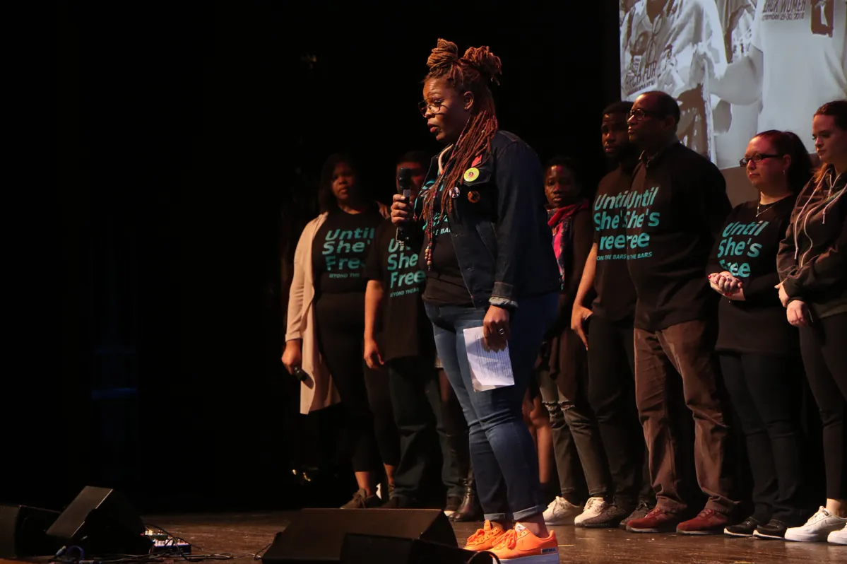 a woman standing on a stage talking into a microphone with people behind her all wearing black shirts that say "Until She's Free"