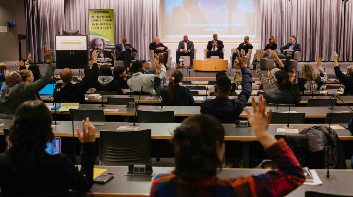 A classroom at CUNY law with students raising their hands and a panel of seven people at the front 