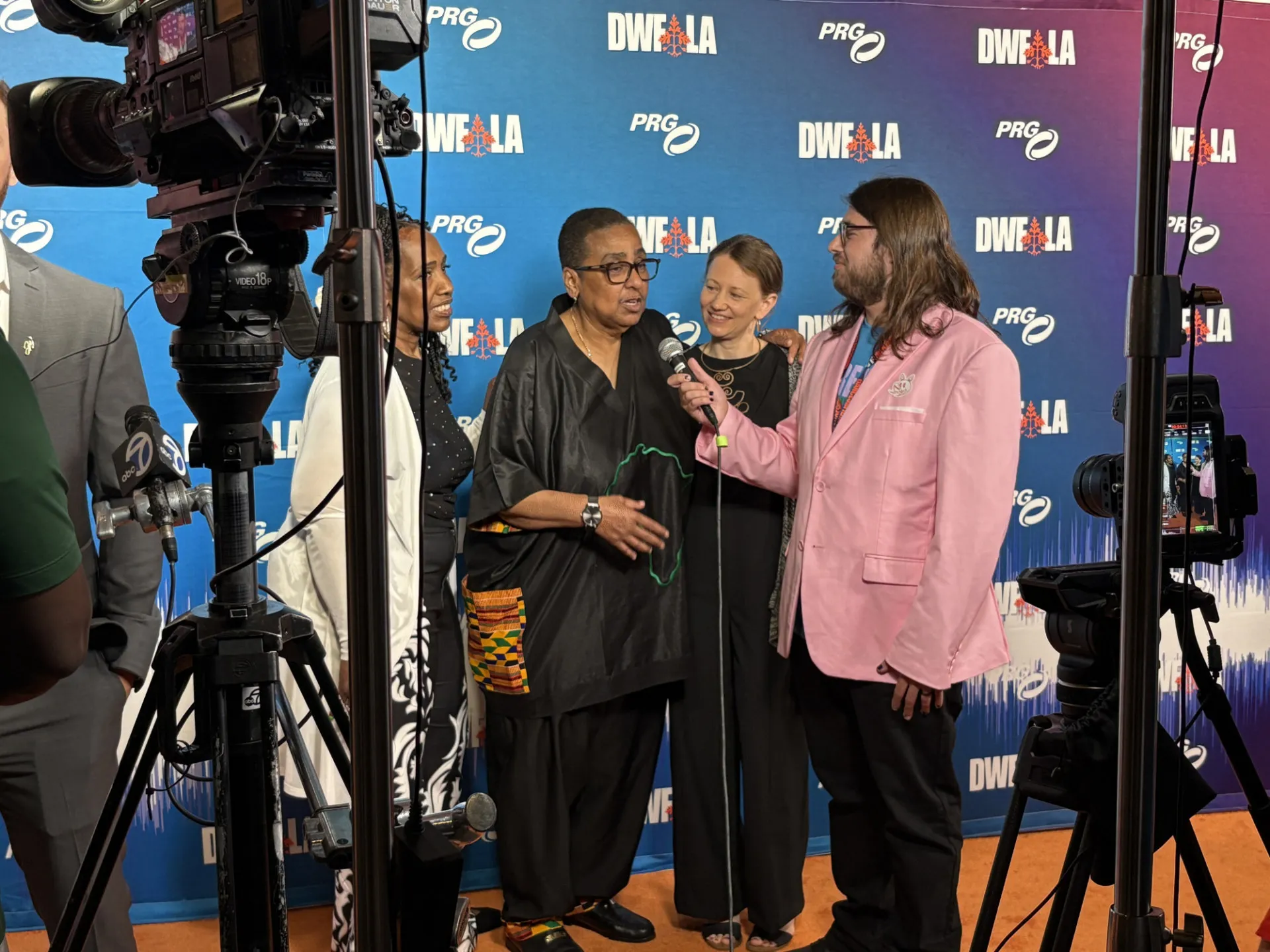 Three women standing in front of a step and repeat speaking into a microphone held by a man in a pink jacket. Cameras are on the side 