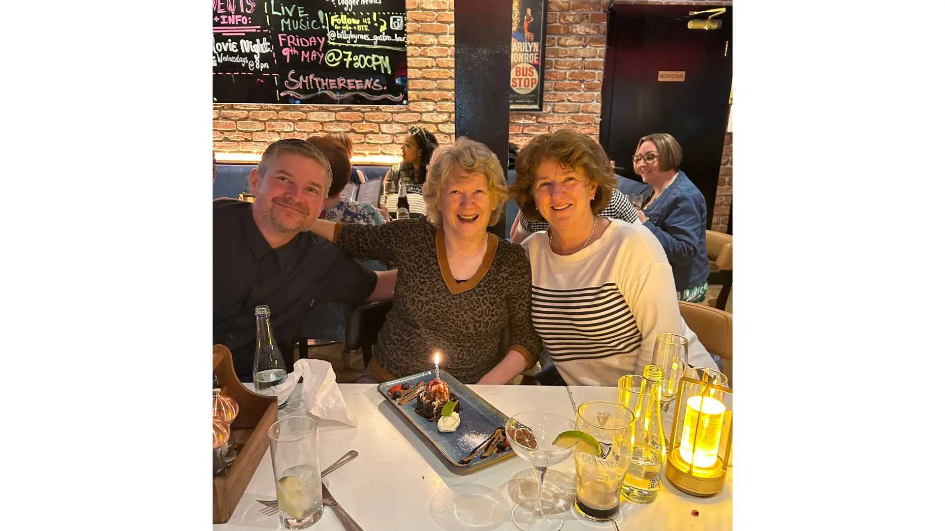 Three people smiling at a restaurant with a cake with a candle on it on the table
