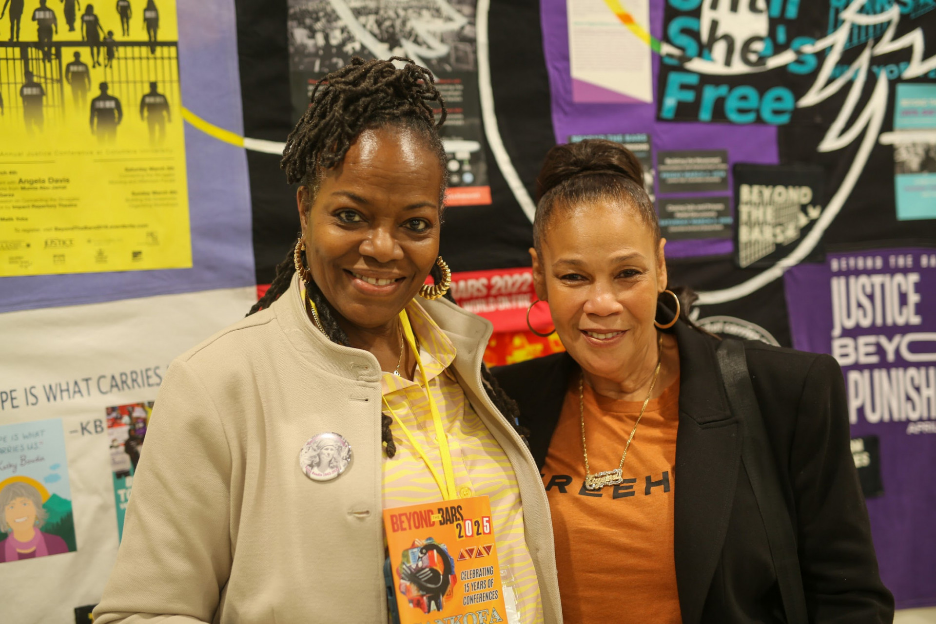 Photo of two women posing together in front of a collage of flyers and photos behind them. One is holding up a program that says Beyond the Bars 2025