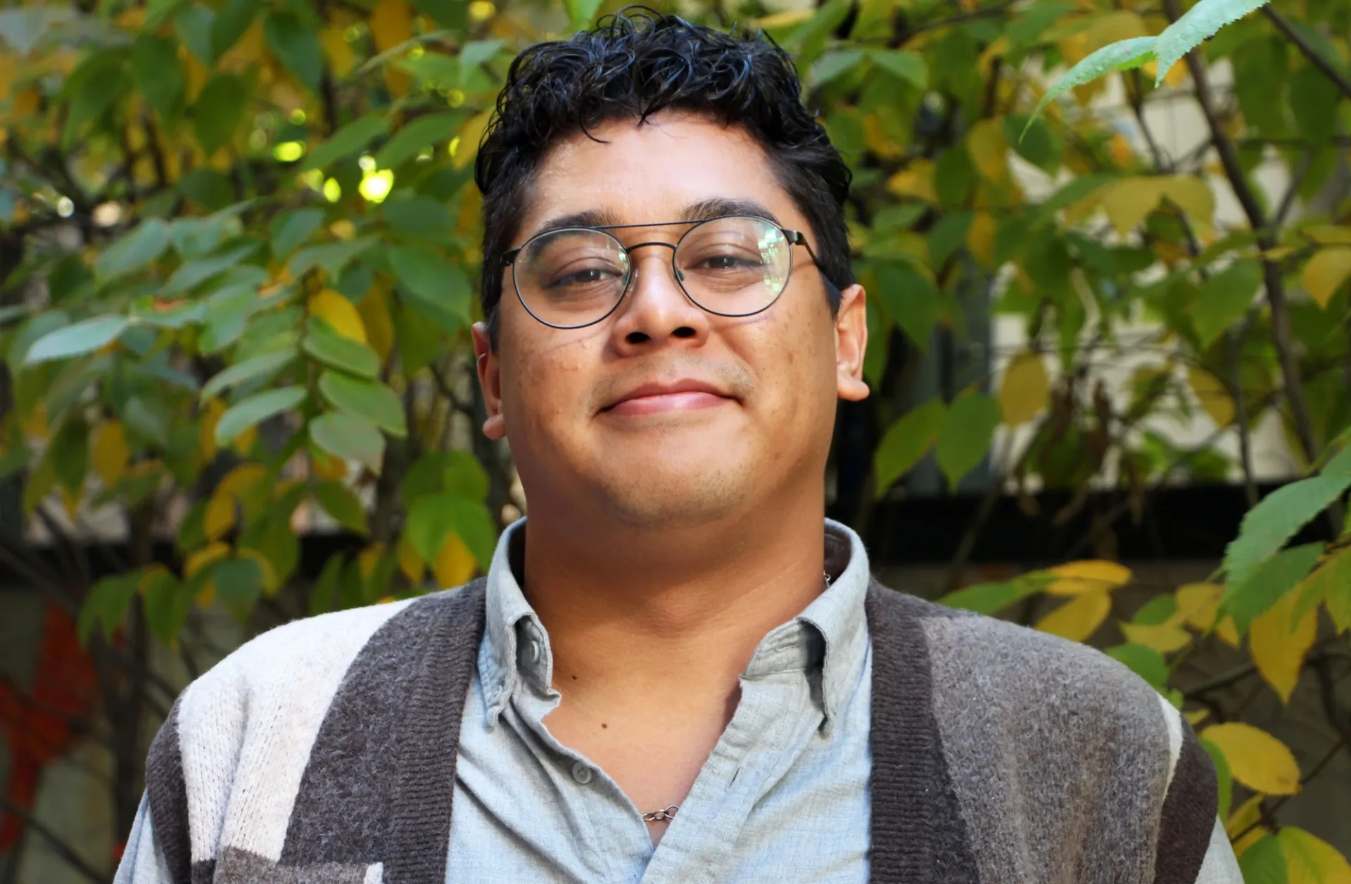Photo of Joe, a man with dark hair and classes and a grey vest in front of some trees