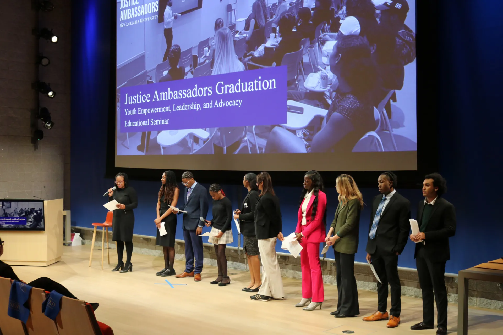 A group of people standing in a line on a stage in front of a screen that says "Justice Ambassadors Graduation"