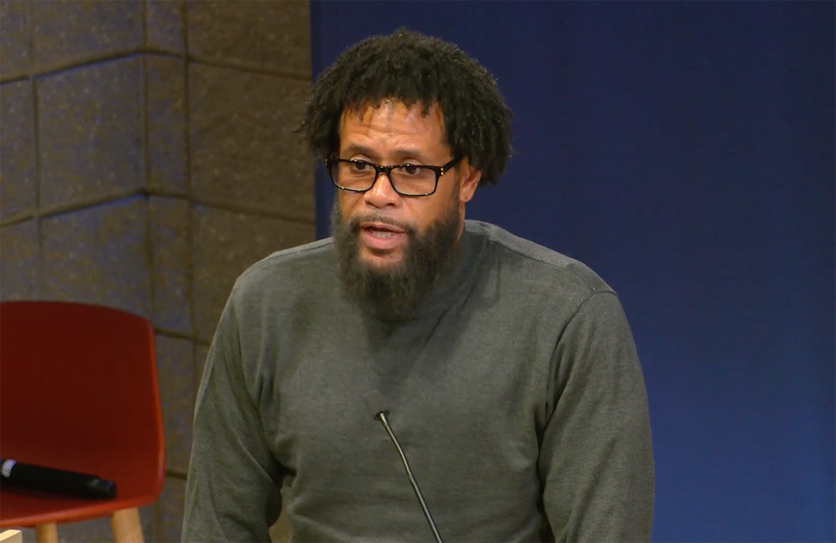 Bill Haney, a Black man with glasses and a beard, standing at a podium with a mic talking