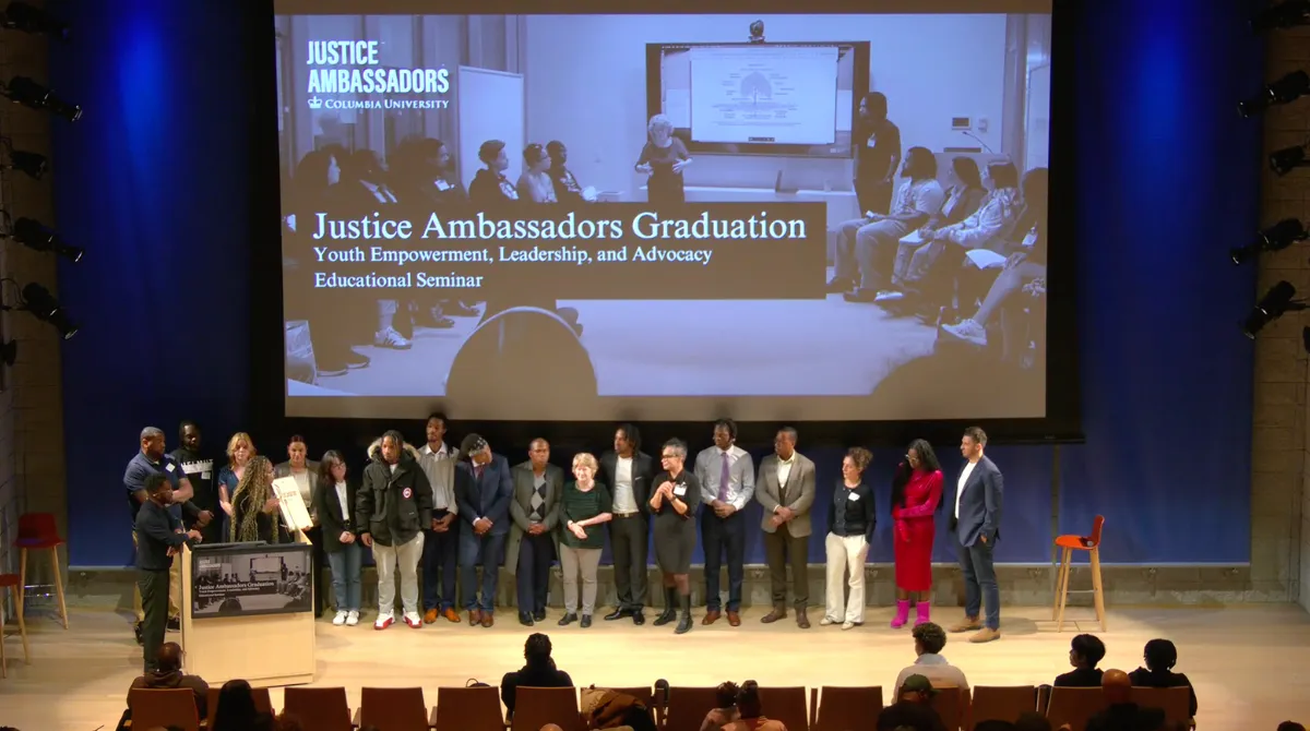 A group of people standing in a line in front of a crowd with a screen in the background that says "Justice Ambassadors Graduation: Youth Empowerment, Leadership, and Advocacy Educational Seminar"