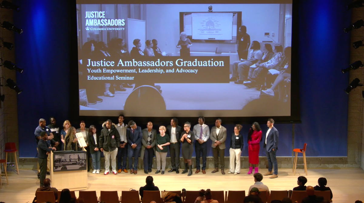 A group of people standing in a line in front of a crowd with a screen in the background that says "Justice Ambassadors Graduation: Youth Empowerment, Leadership, and Advocacy Educational Seminar"
