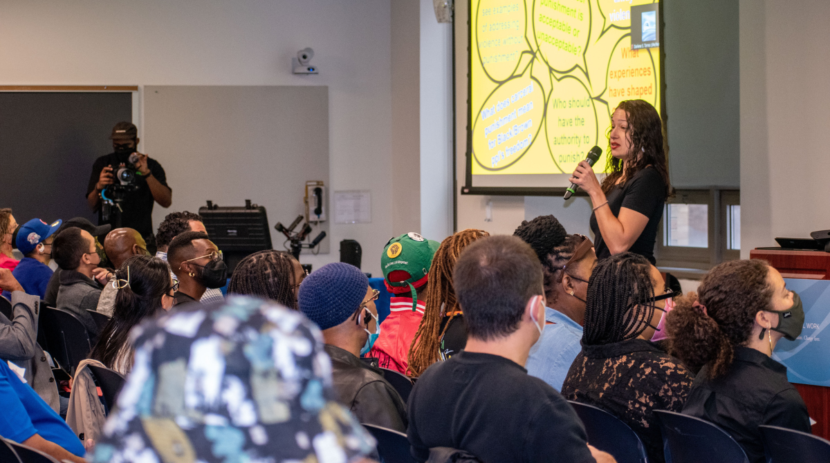 Woman standing in front of a packed classroom presenting with a screen behind her with a yellow image with quote bubbles