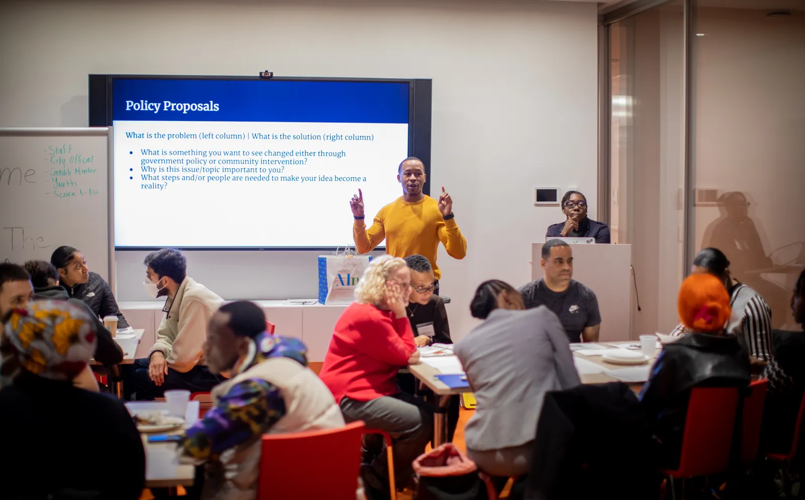 A man standing in front of a group of people presenting with a screen behind him that says "Policy Proposals"