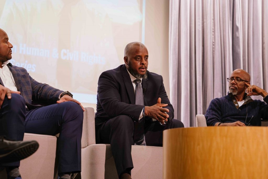 A Black man in a suit sitting in a chair with his hands together speaking to a crowd with two other Black men sitting on each side of him
