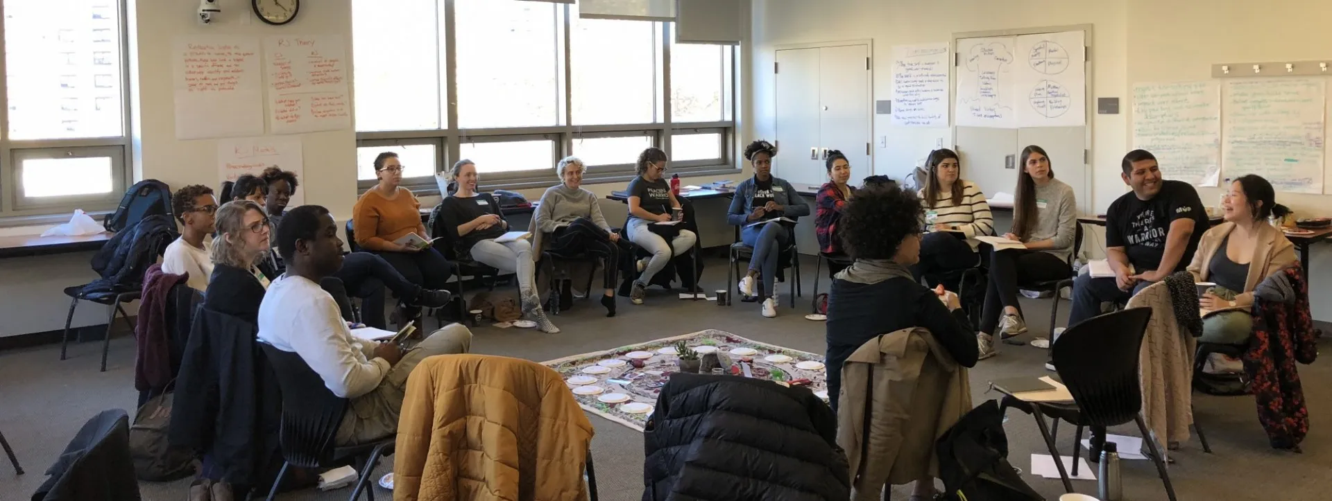 group of people sitting in a circle around a center piece of a rug and items from the group