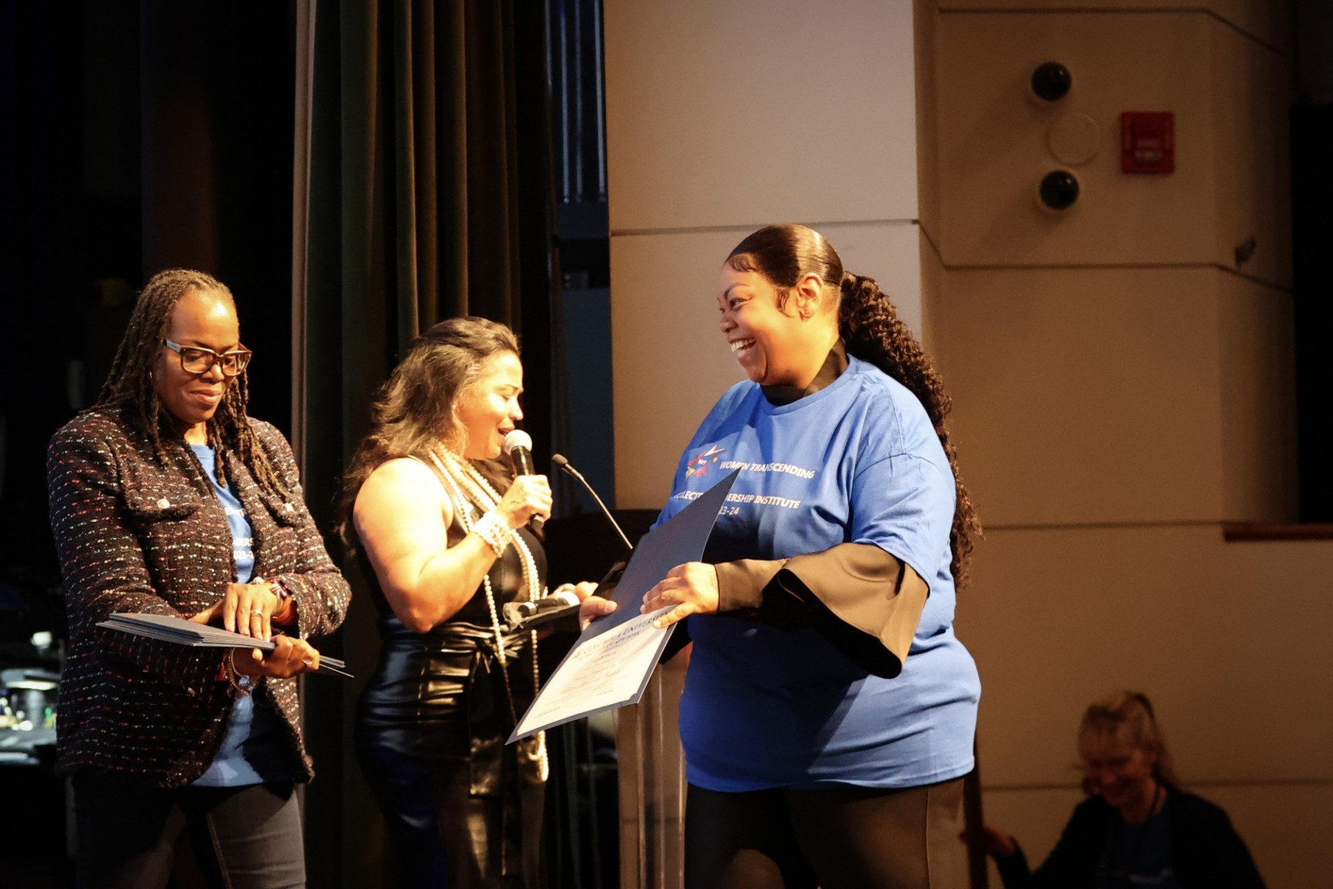 A woman in a blue shirt walking across a stage smiling and receiving a certificate from another woman