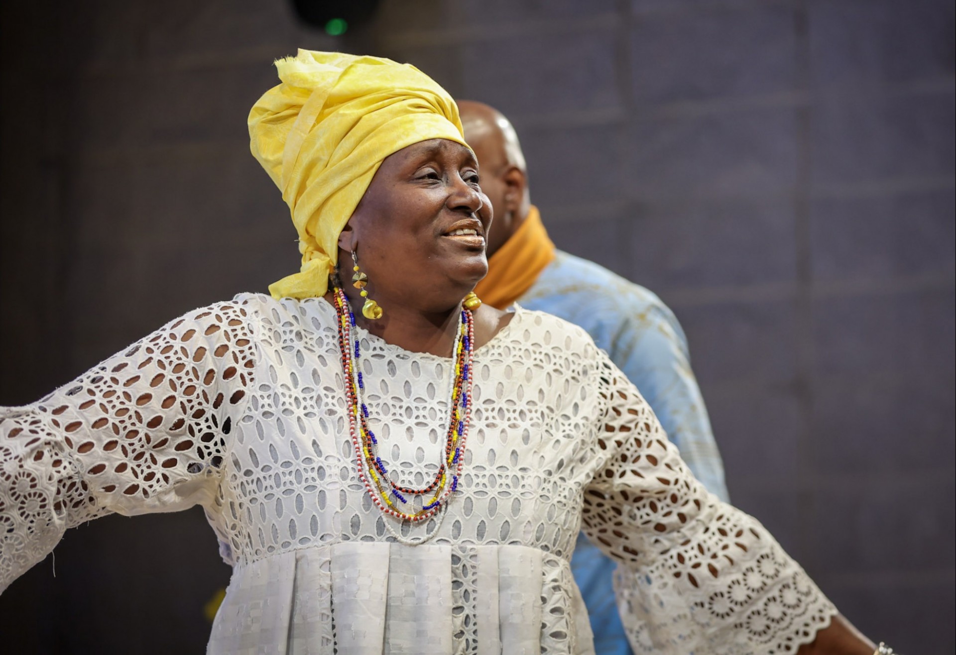A picture of a Black woman with a white dress and yellow wrap around her head holding her arms out and smiling