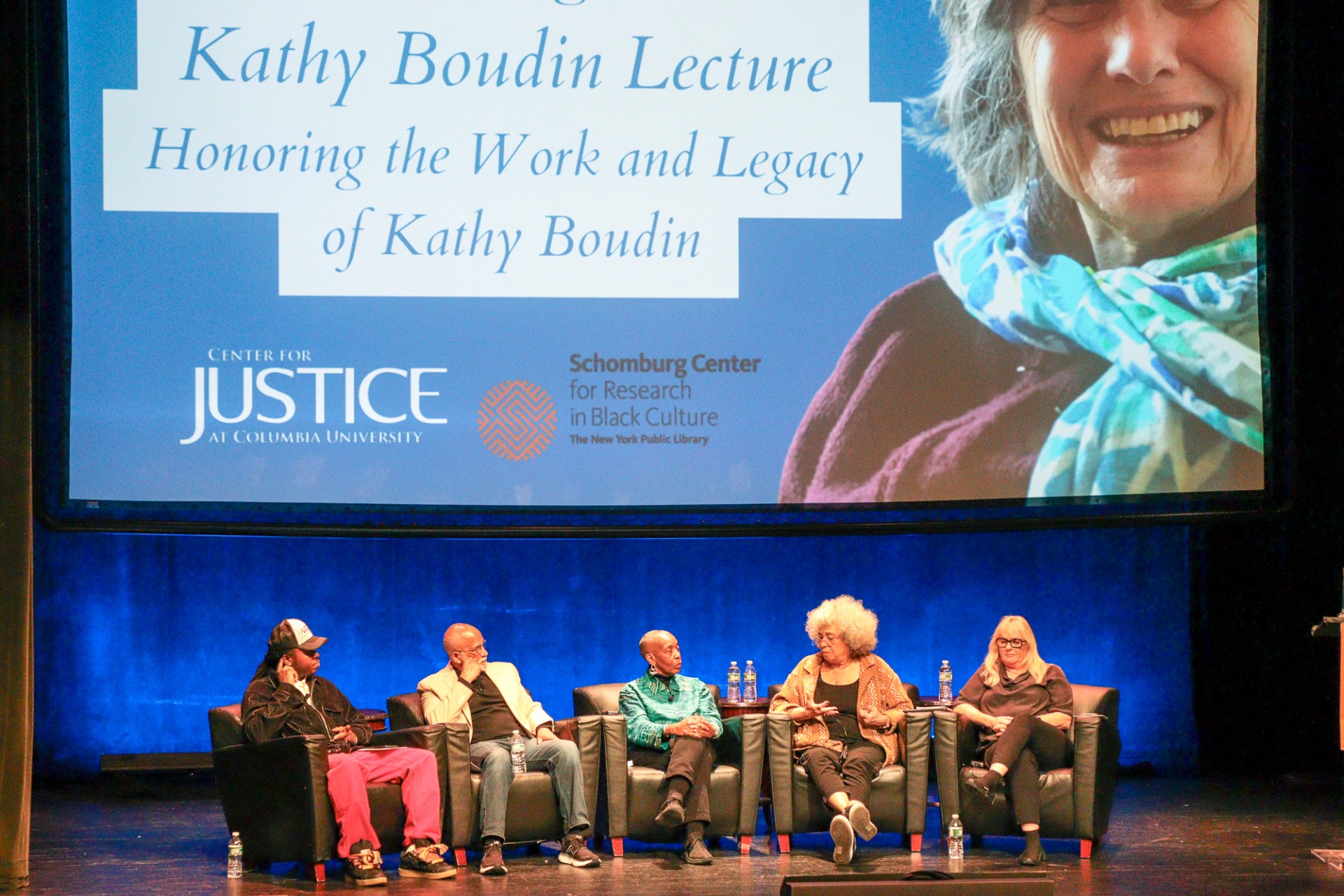 Picture of the panelists sitting on chairs on stage with an image of Kathy on the screen behind them