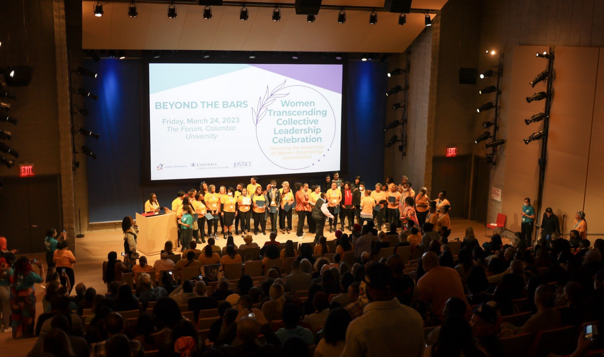 wide shot of the room/audience with a group on stage in yellow tshirts and a screen in the background that says "Women Transcending Collective Leadership Celebration: