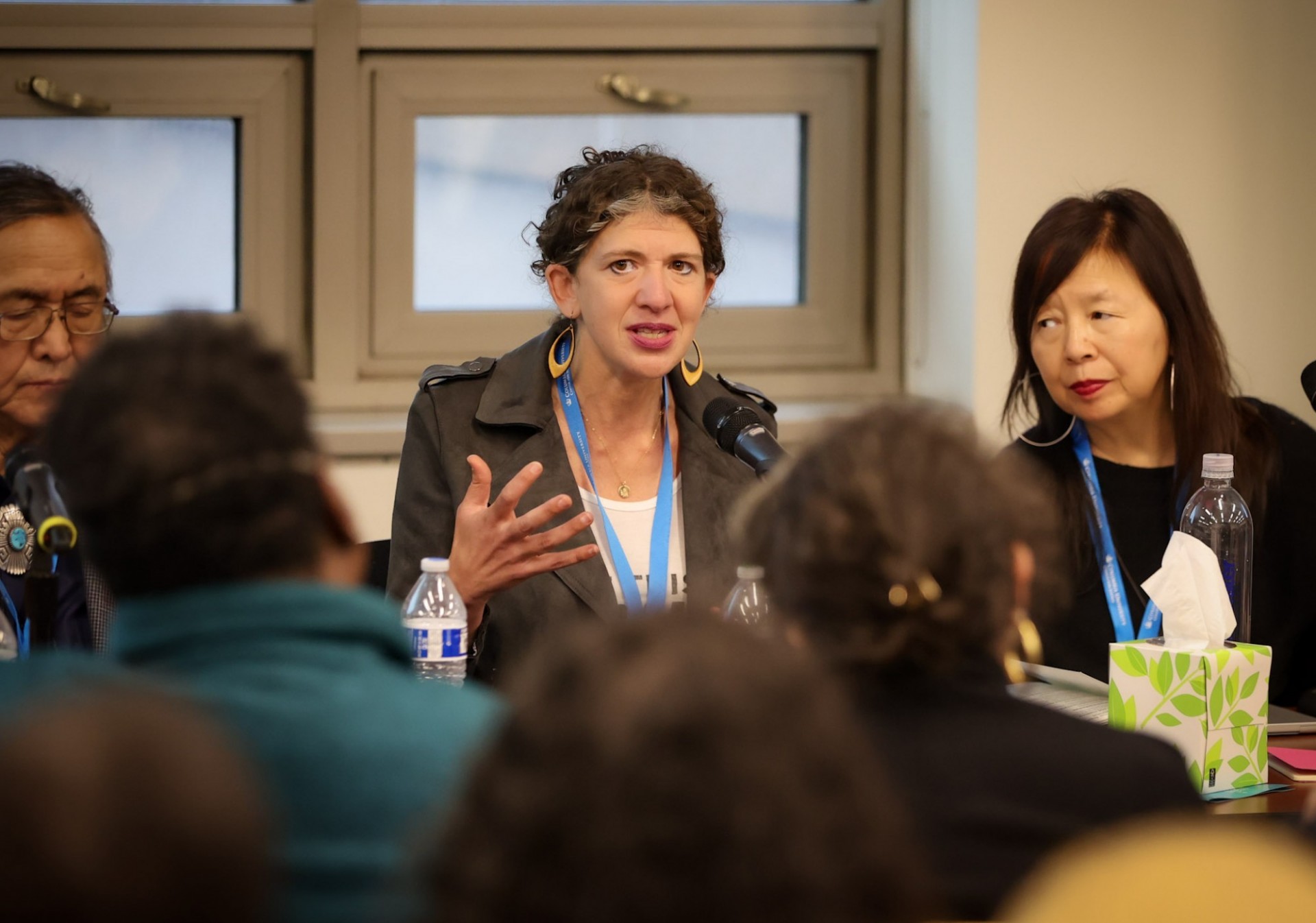 picture of a woman speaking at a table in front of a microphone and audience with two panelists on each side of her