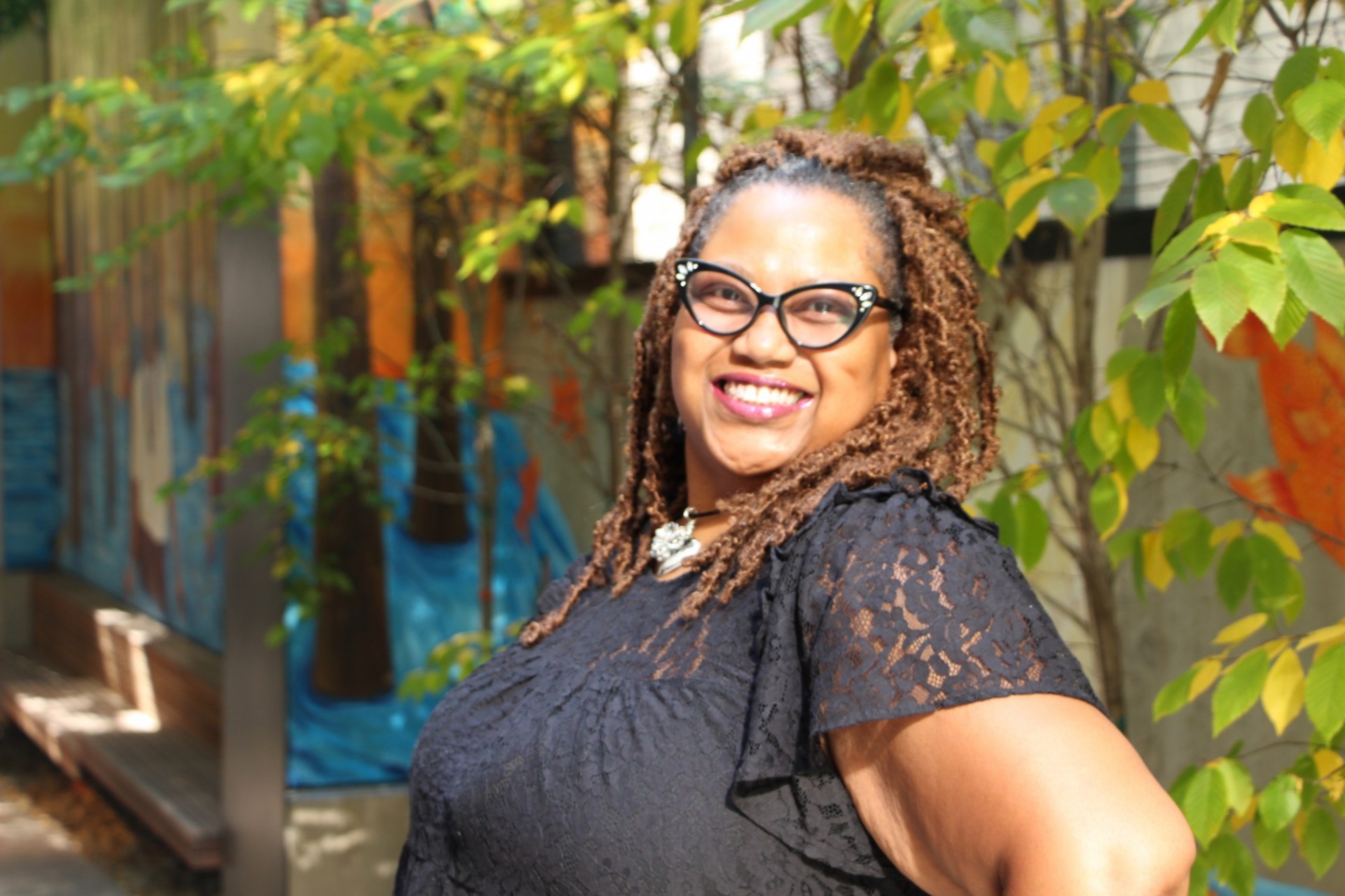 photo of Crystal, a Black woman smiling in a black shirt and glasses, in front of some trees 