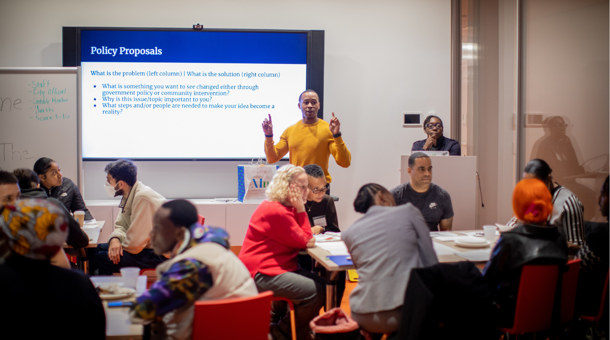 group of people seated around tables listening to a guy speaking at the front with a screen behind him that says "Policy Proposals"