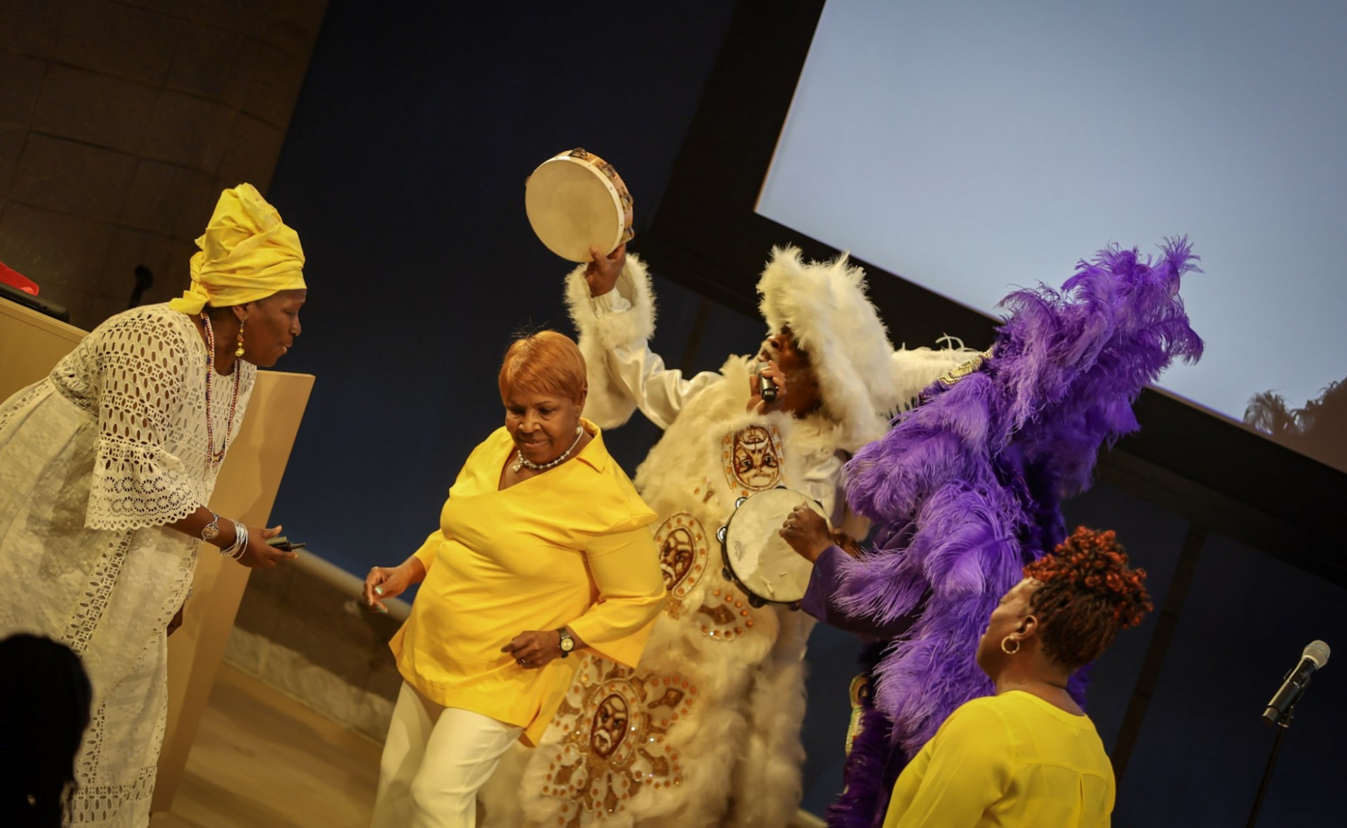 A group of people dancing on stage, some dressed in purple and white feathered headdresses. One is holding a tamborine. 