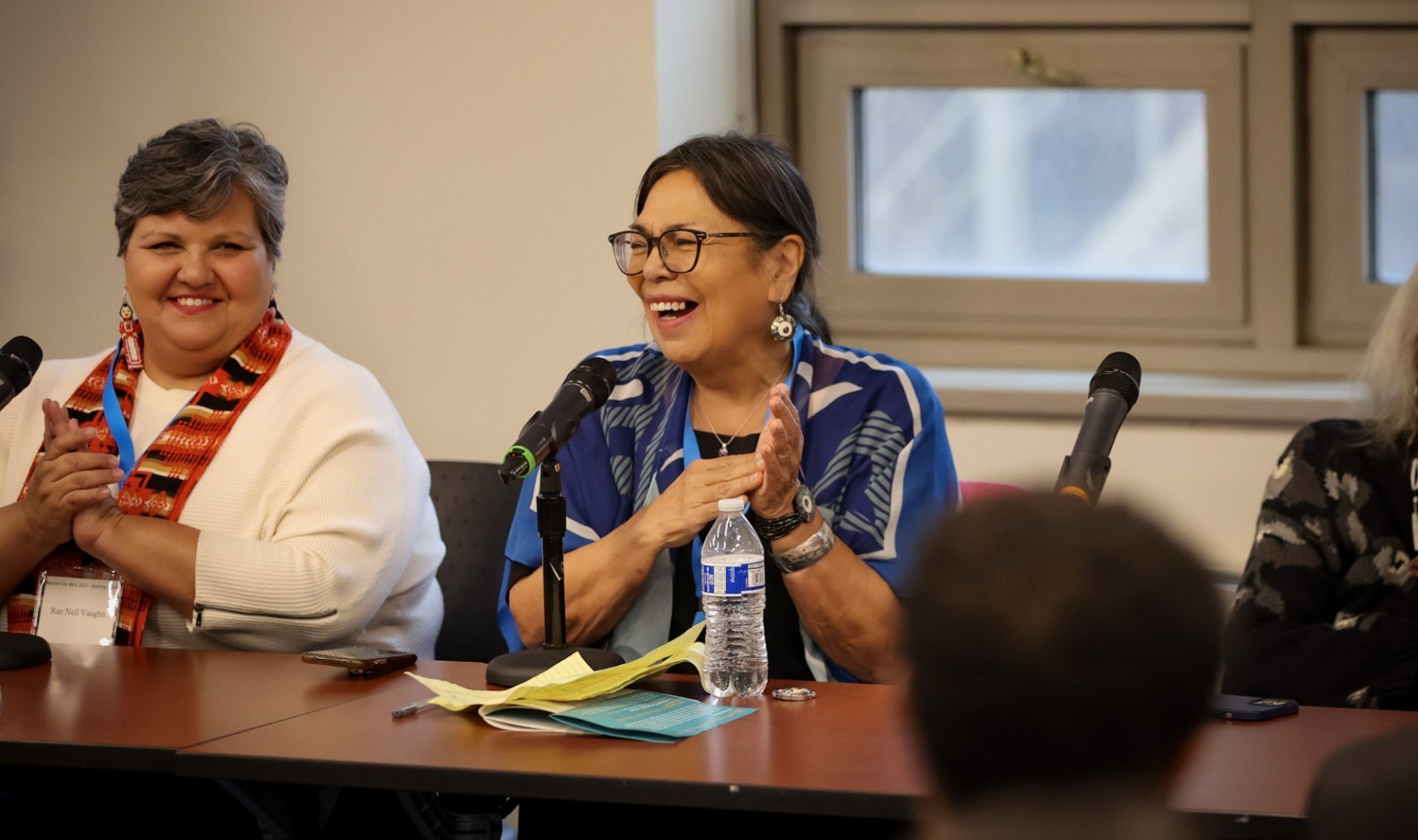 Two women on a panel at a table smiling with microphones in front of them