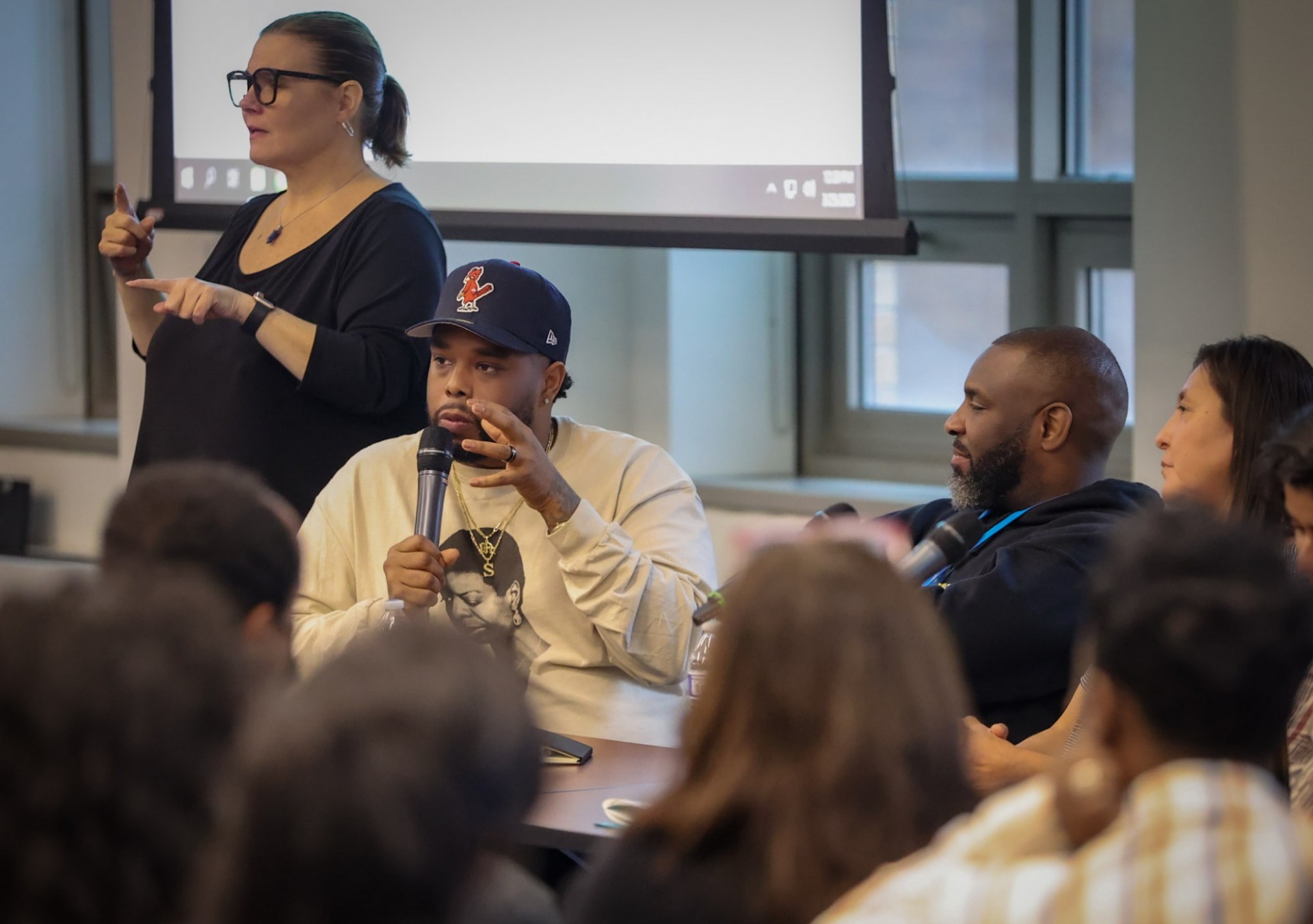 A Black man holding a microphone sitting at table in front of an audience in a classroom with an interpreter on his left and the other panelist on his right