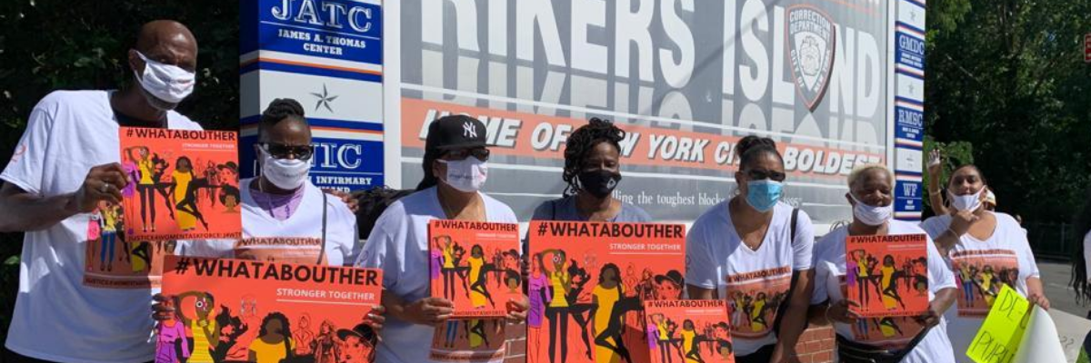 Advocates holding up signs that say What About Her and Stronger Together outside the Rikers Island sign