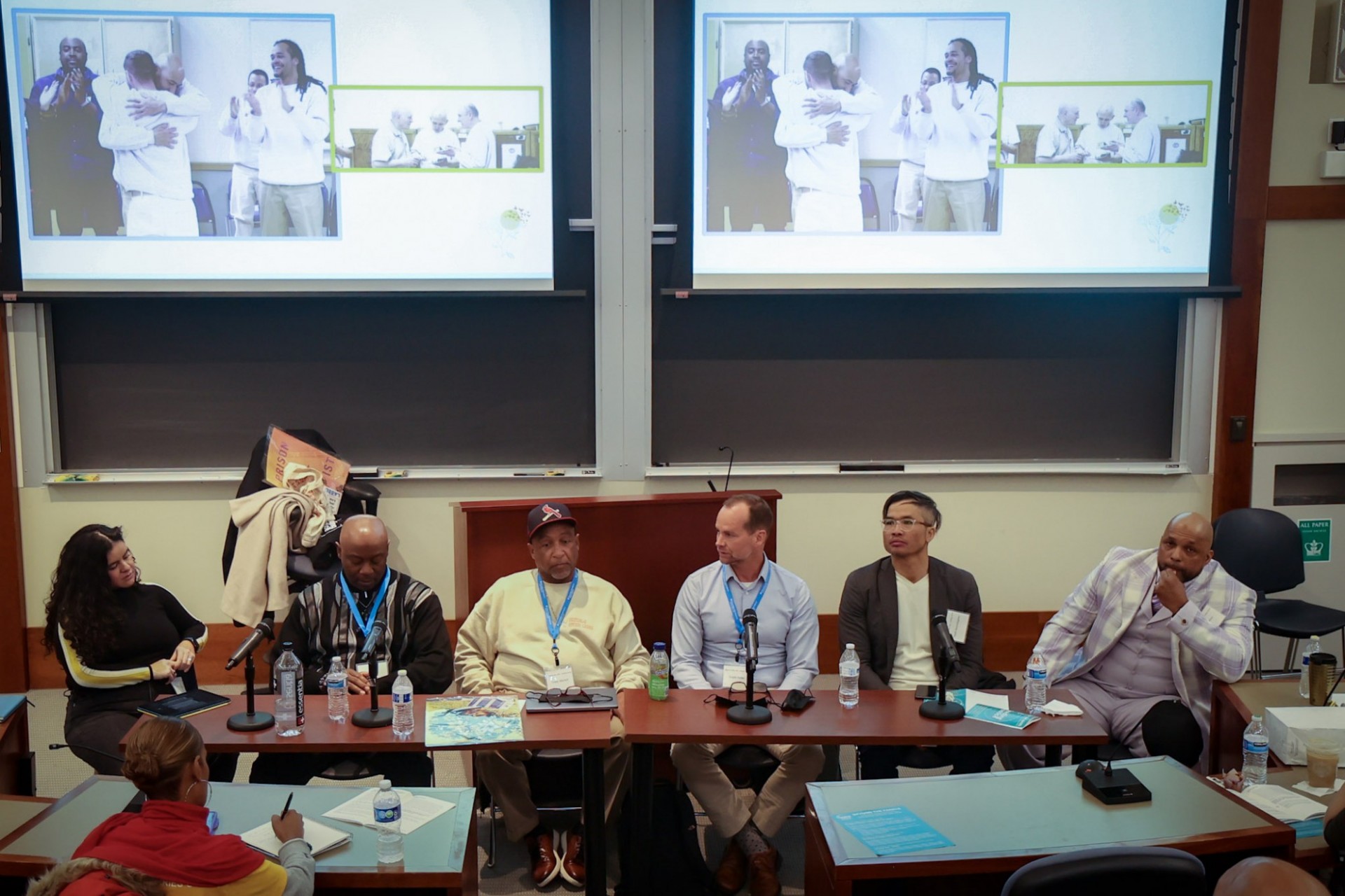 A panel of people in a classroom conducting a workshop with screens behind them that are Zooming in some currently incarcerated men to participate