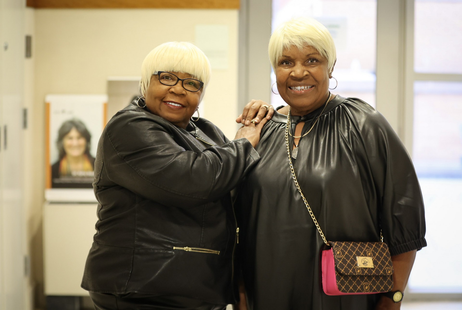 Two Black women, wearing all black, with short light hair standing next to each other posing and smiling