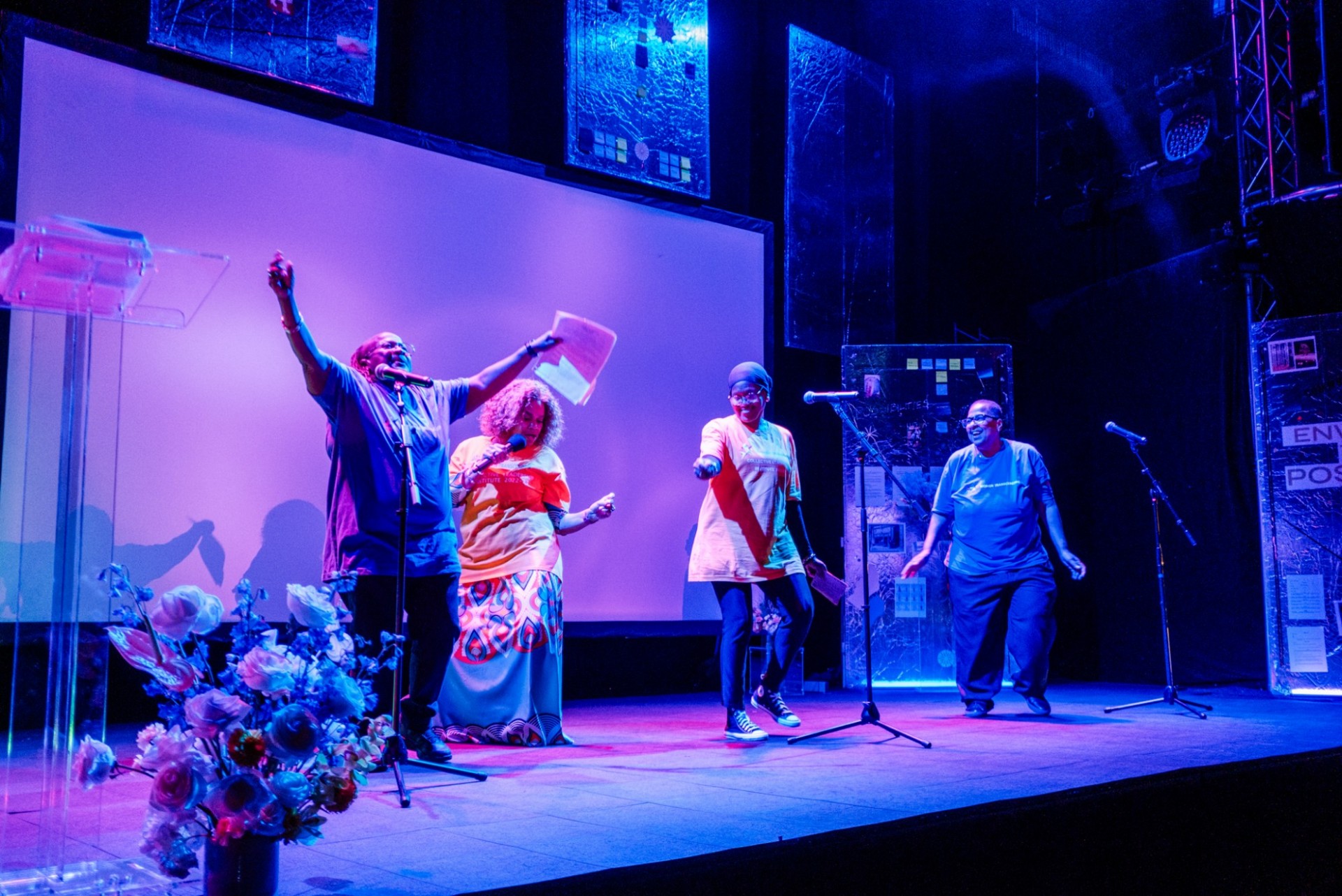 Four women on a stage with purple lighting, performing a spoken word