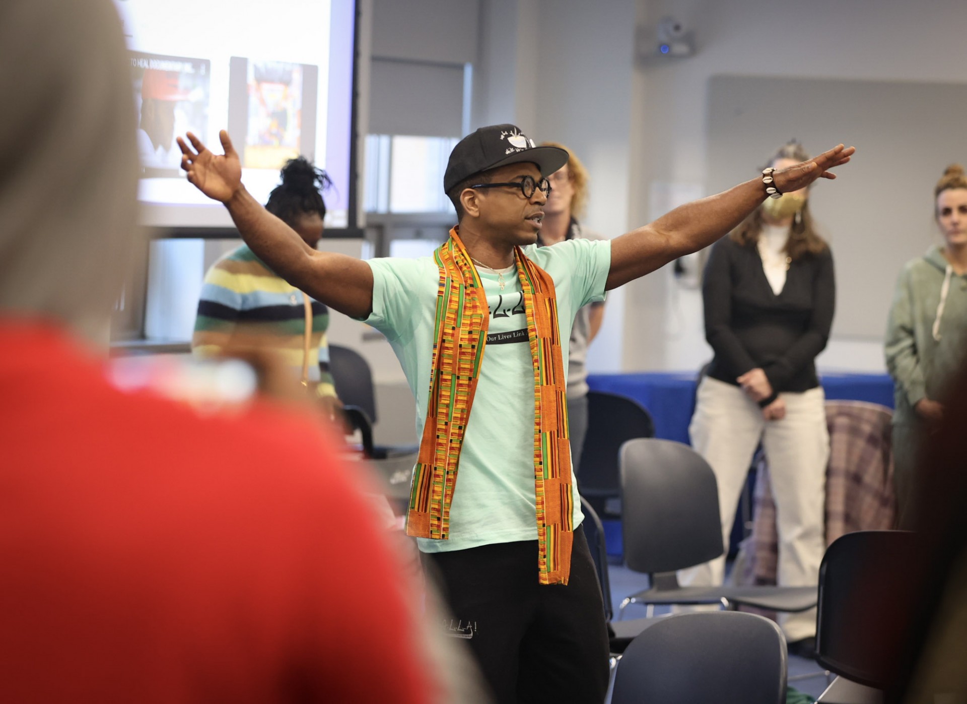 A Black man with a colorful scarf on standing in the middle of a circle of people with his arms lifted